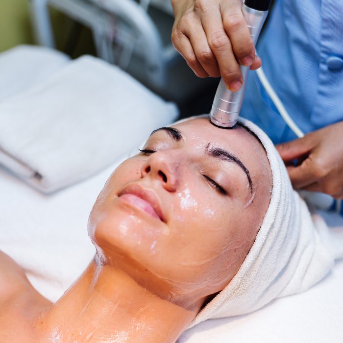 Woman lying with eyes closed as a skincare professional uses a silver handheld device on her forehead during a facial treatment.